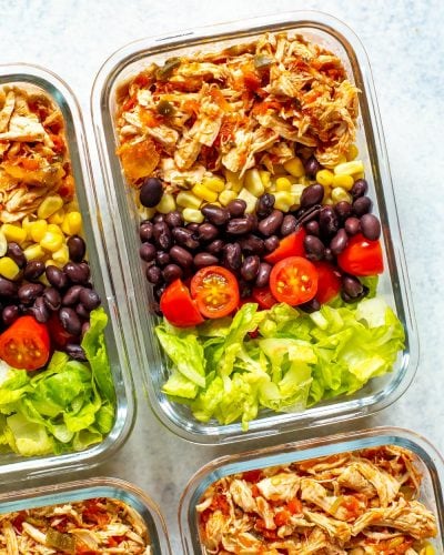 A close-up of four meal prep containers, each containing crockpot salsa chicken, corn, black beans, cherry tomatoes and lettuce.