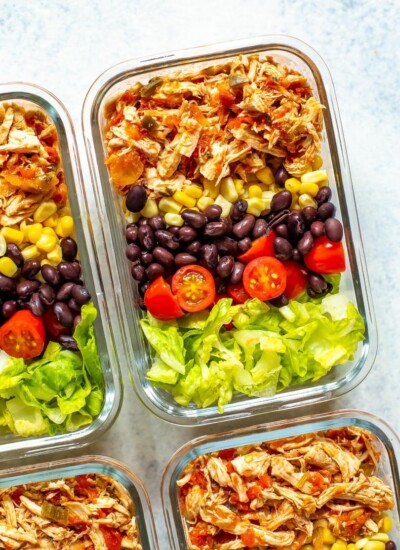 A close-up of four meal prep containers, each containing crockpot salsa chicken, corn, black beans, cherry tomatoes and lettuce.