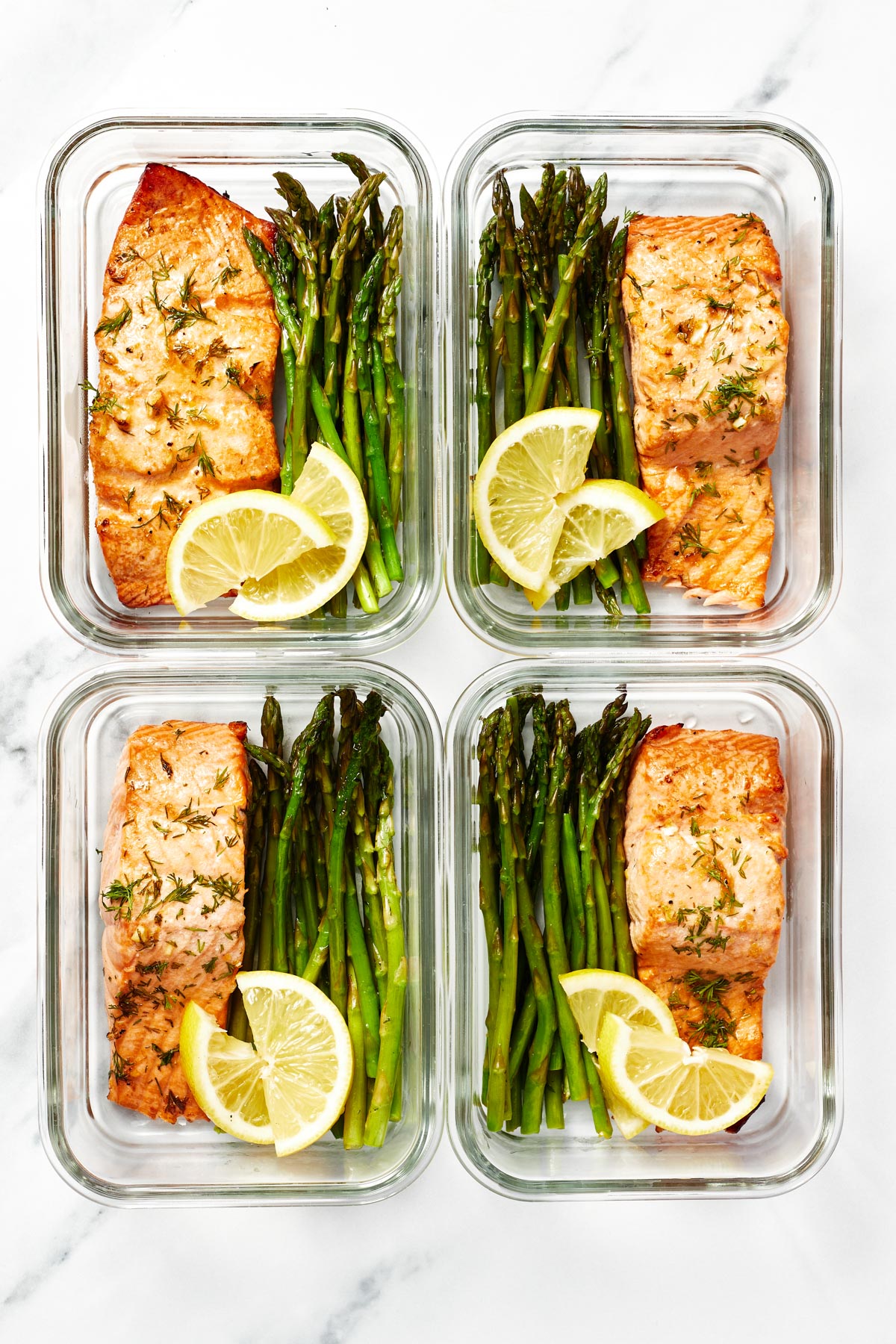 Four meal prep containers, each with a filet of air fryer salmon, asparagus and two lemon slices.