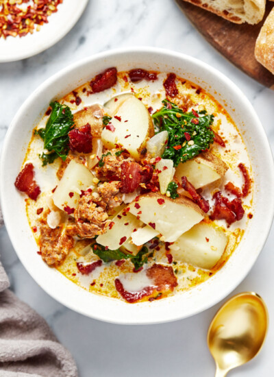 A bowl of zuppa toscana with red chili flakes and bread on the side.