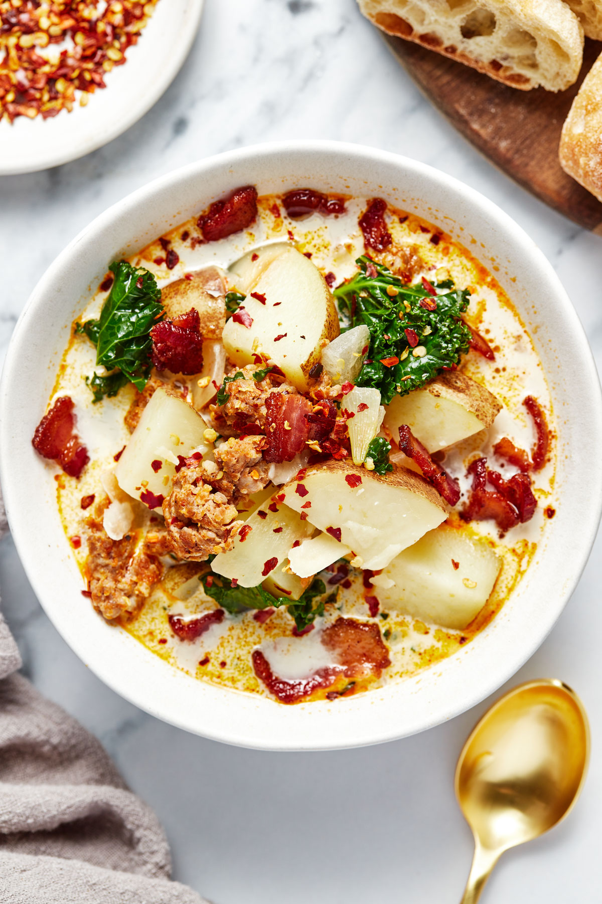 A bowl of zuppa toscana with red chili flakes and bread on the side.