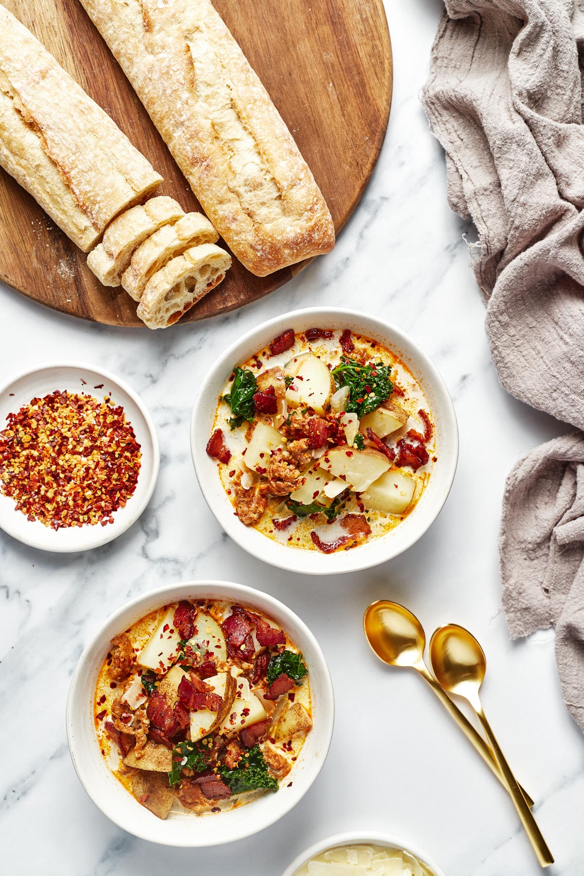 An overhead shot of two bowls of zuppa toscana with two sliced baguettes and a bowl red chili flakes to the side. 