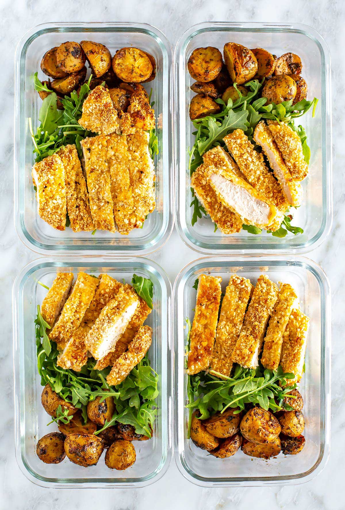 Four meal prep containers, each with a bed of arugula, lemon dill potatoes and a sliced chicken cutlet on top.