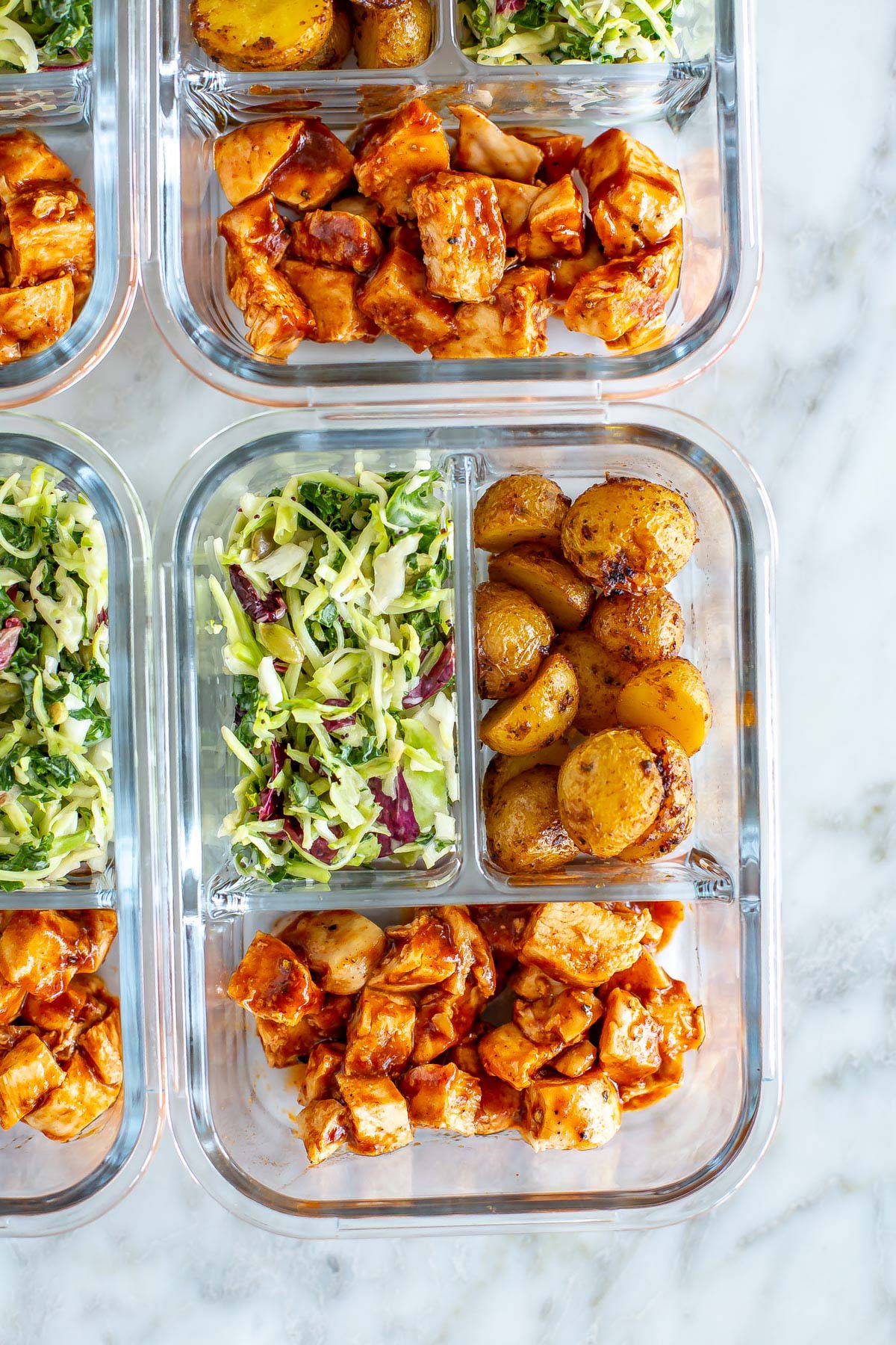 A close-up of a meal prep container with roasted baby potatoes, kale salad and baked BBQ chicken inside.