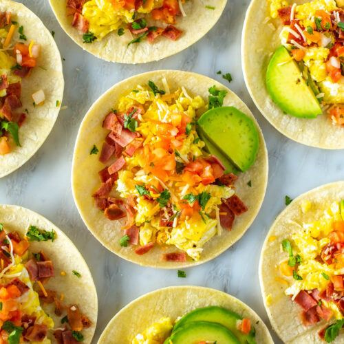 An overhead shot of breakfast tacos topped with eggs, bacon, pice de gallo, avocado and cheese.
