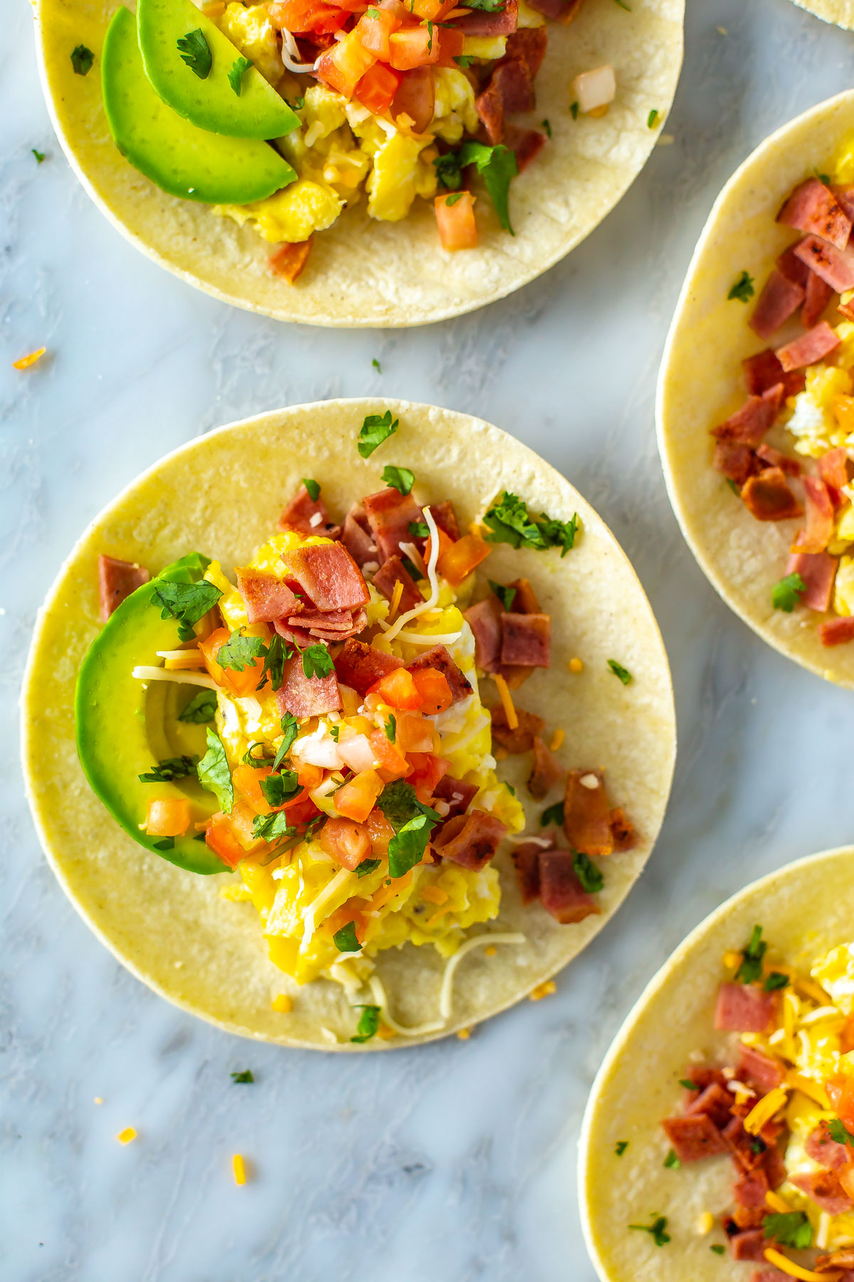 A close-up overhead shot of breakfast tacos topped with eggs, bacon, pice de gallo, avocado and cheese.