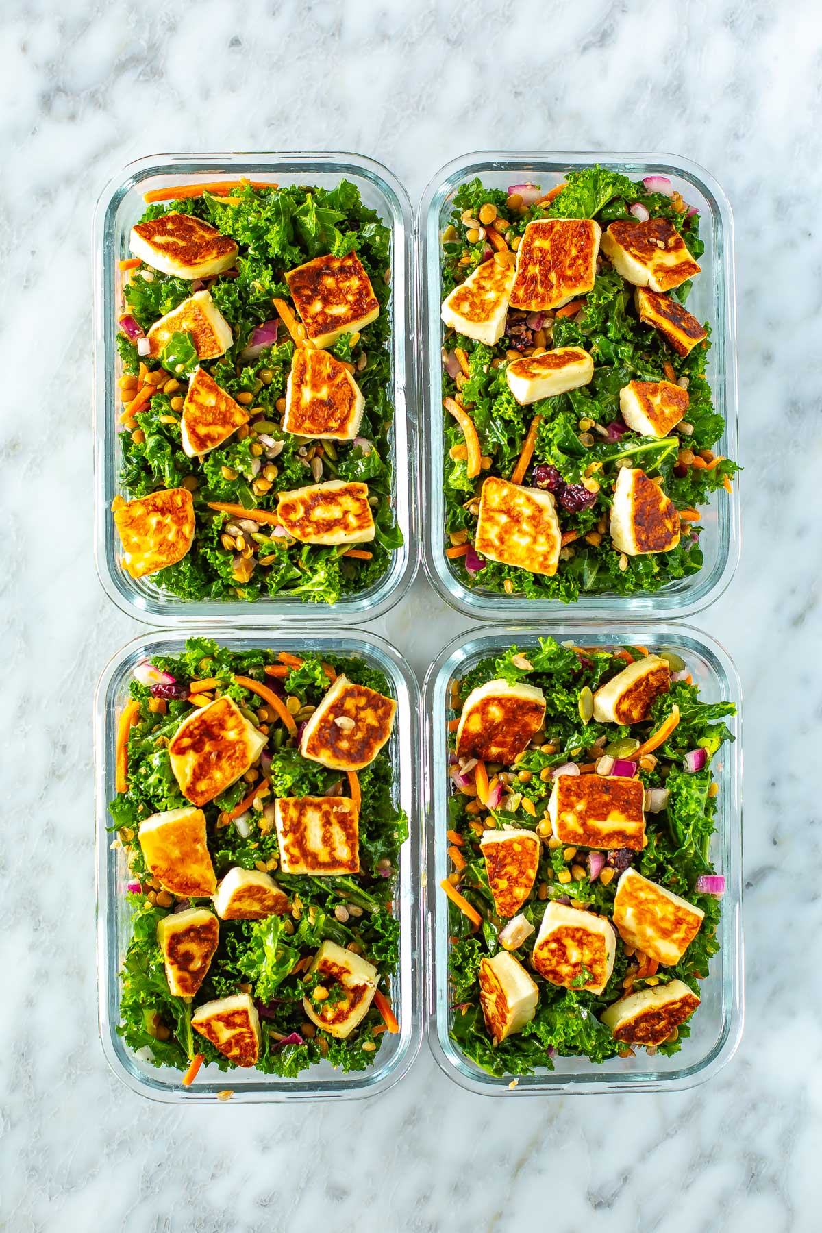 Four meal prep containers, each filled with kale salad topped with halloumi cheese.