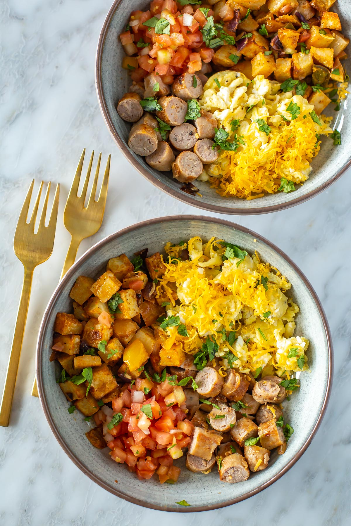 Two breakfast bowls filled with roasted veggies, scrambled eggs and cooked sausage.