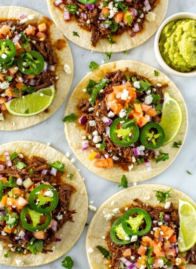 Five shredded beef tacos, each with an assortment of toppings on top.