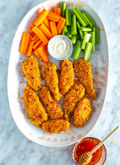 A platter with a bunch of air fryer hot honey chicken tenders, celery, carrots and ranch dressing placed next to a bowl of hot honey.