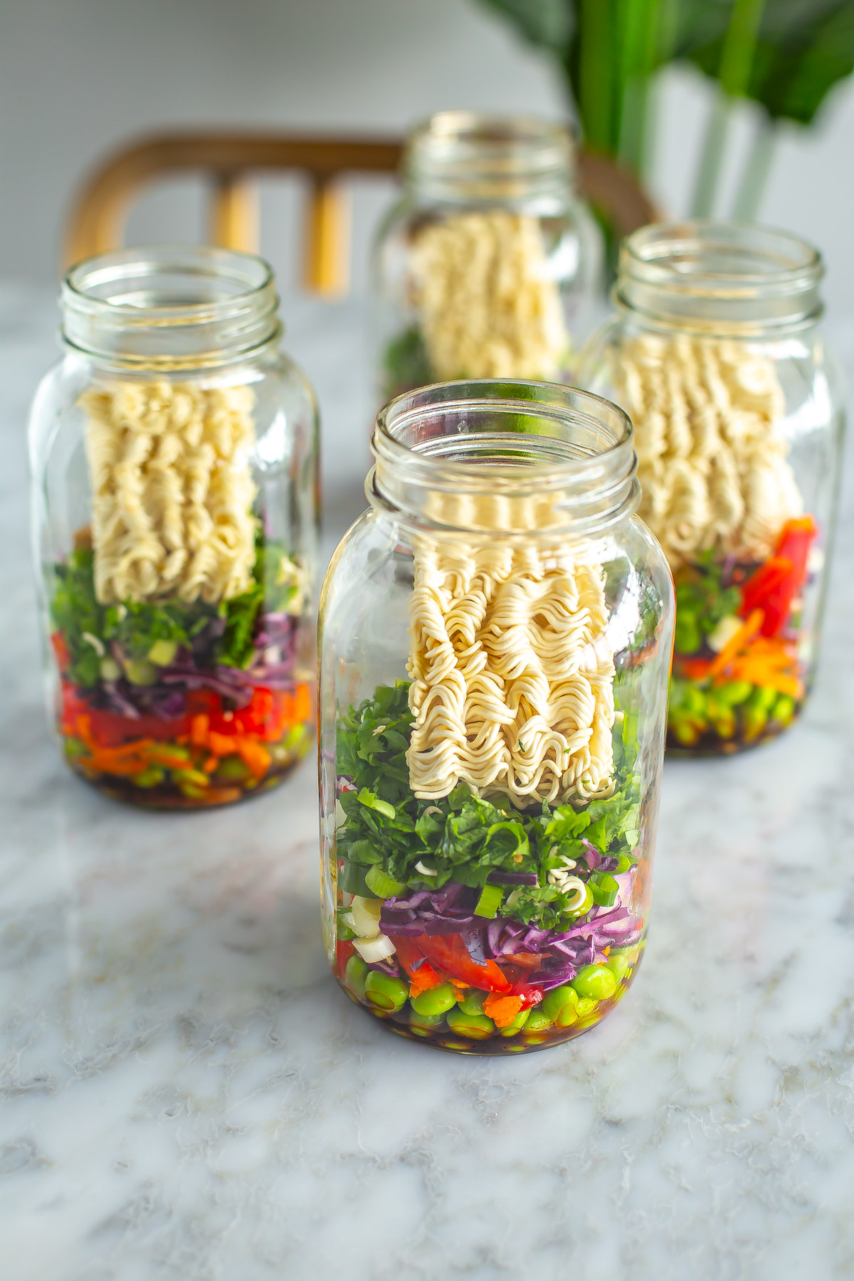 Four mason jars, each filled with instant noodles, vegetables and herbs.