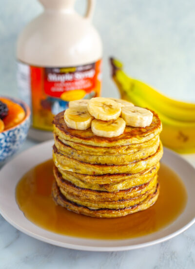 A stack of banana egg pancakes with extra banana slices and maple syrup on top.