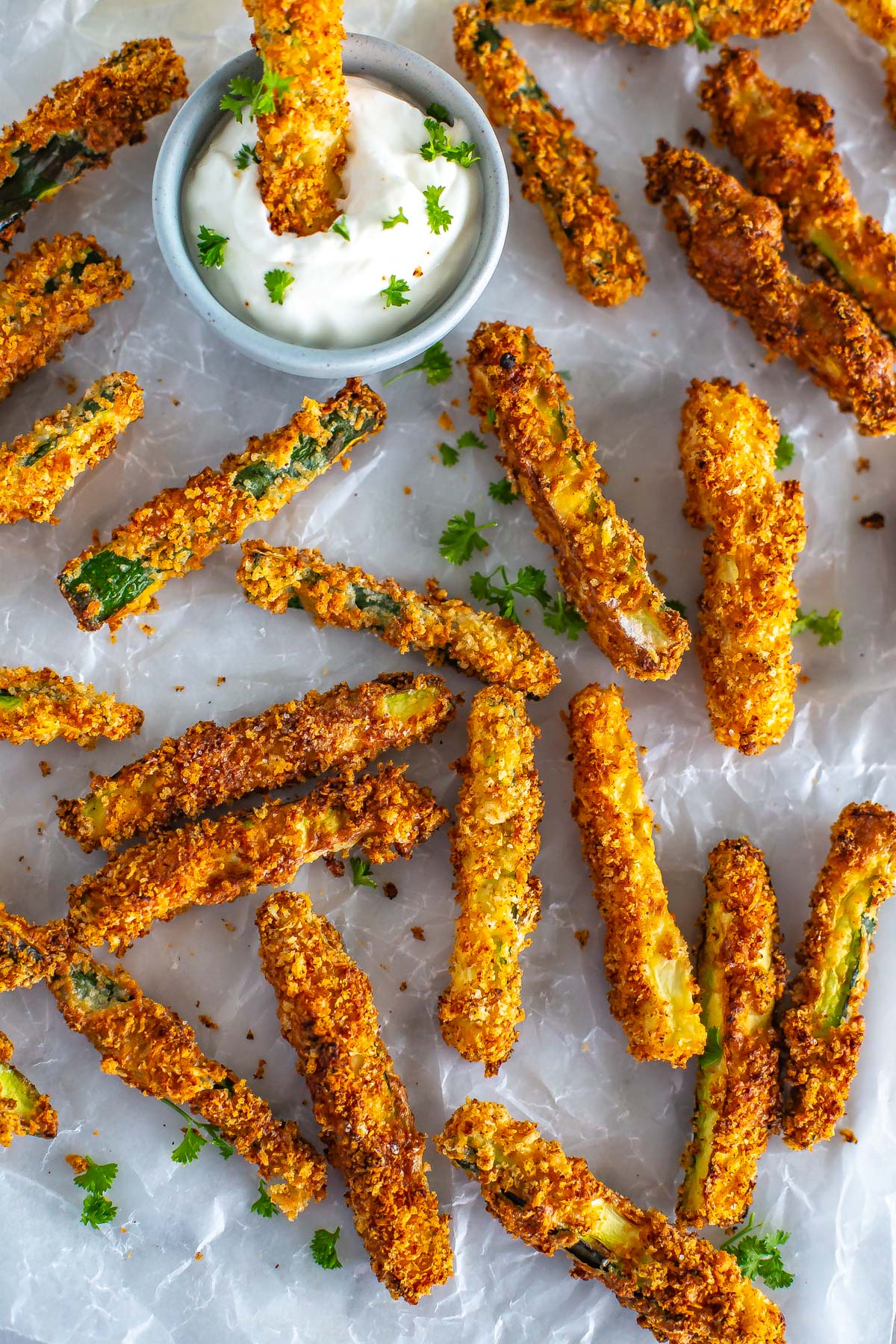 Multiple air fryer zucchini fries on a sheet of parchment paper, with one being dipped into garlic aioli.