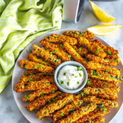 A plate of air fryer zucchini fries with garlic aioli in front of an air fryer.