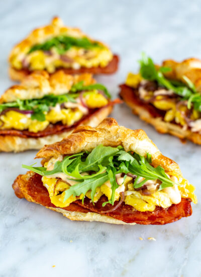 Four croissant breakfast sandwiches with bacon, scrambled eggs and arugula inside them.
