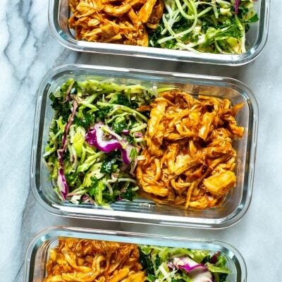 Three meal prep containers, each containing shredded crockpot BBQ chicken and kale slaw.