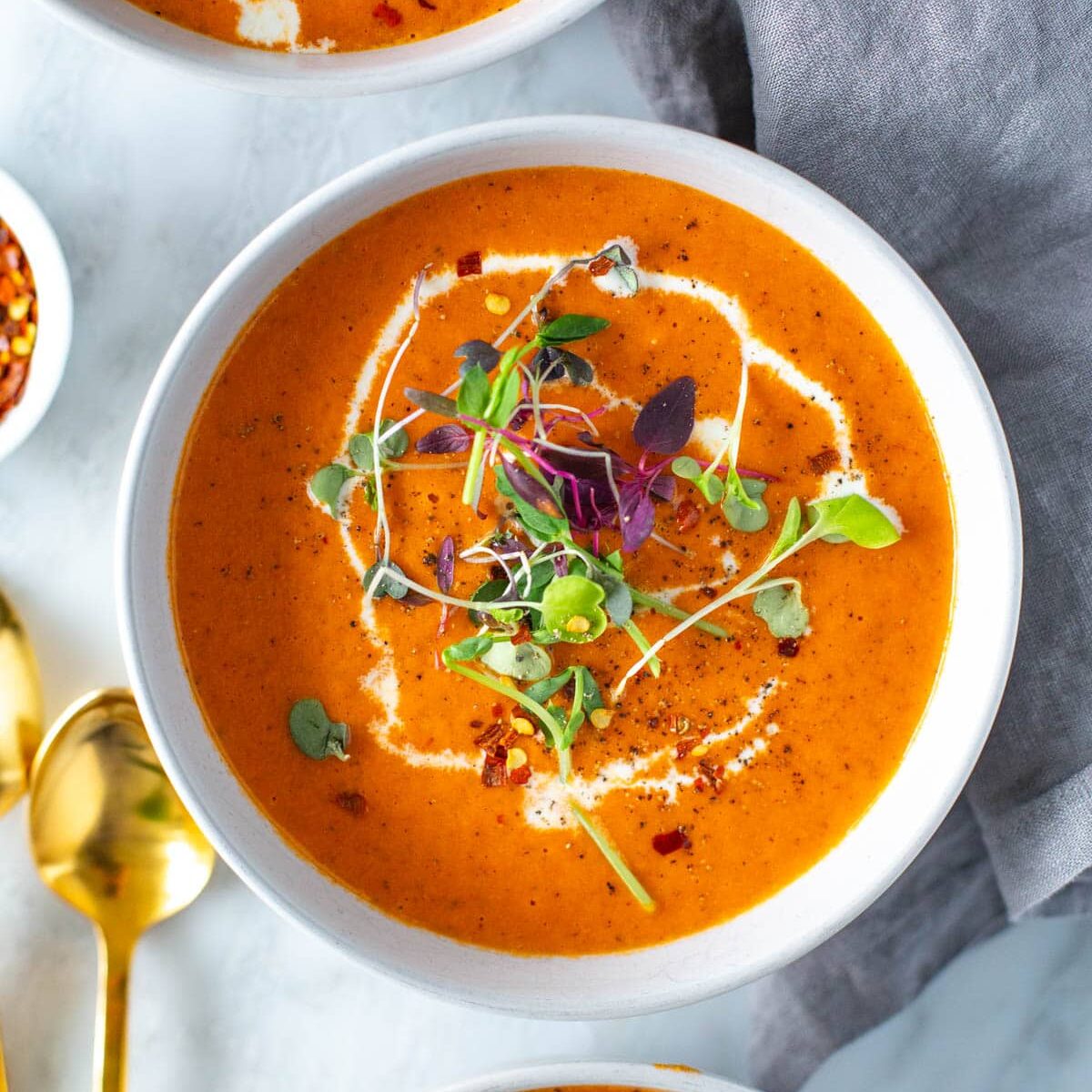 A close-up of a bowl of roasted red pepper soup topped with heavy cream and microgreens.